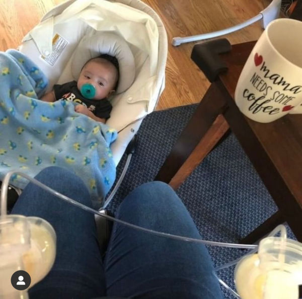baby with a pacifier in mouth on floor next to mom, who is pumping and drinking coffee