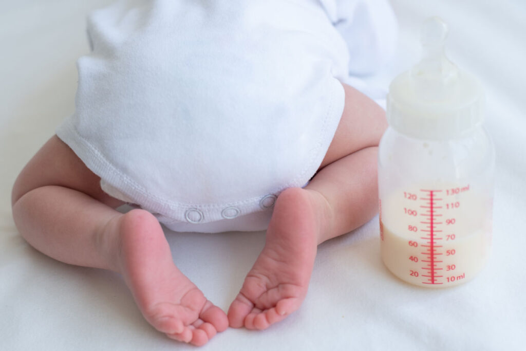 Baby wearing white onesie next to half full bottle of milk