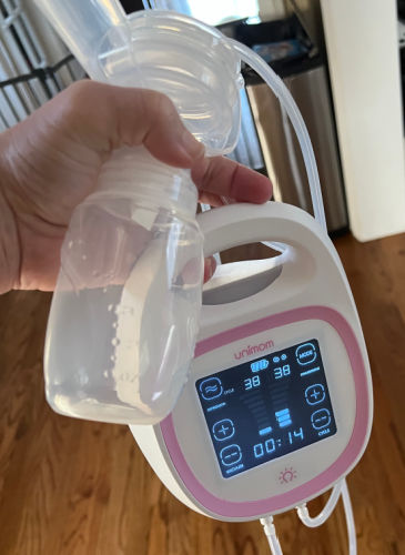 woman holding Unimom Opera breast pump in one hand along with one set of pump parts. The pump is running and the timer is at 14 seconds. The hardwood floor is in the background.